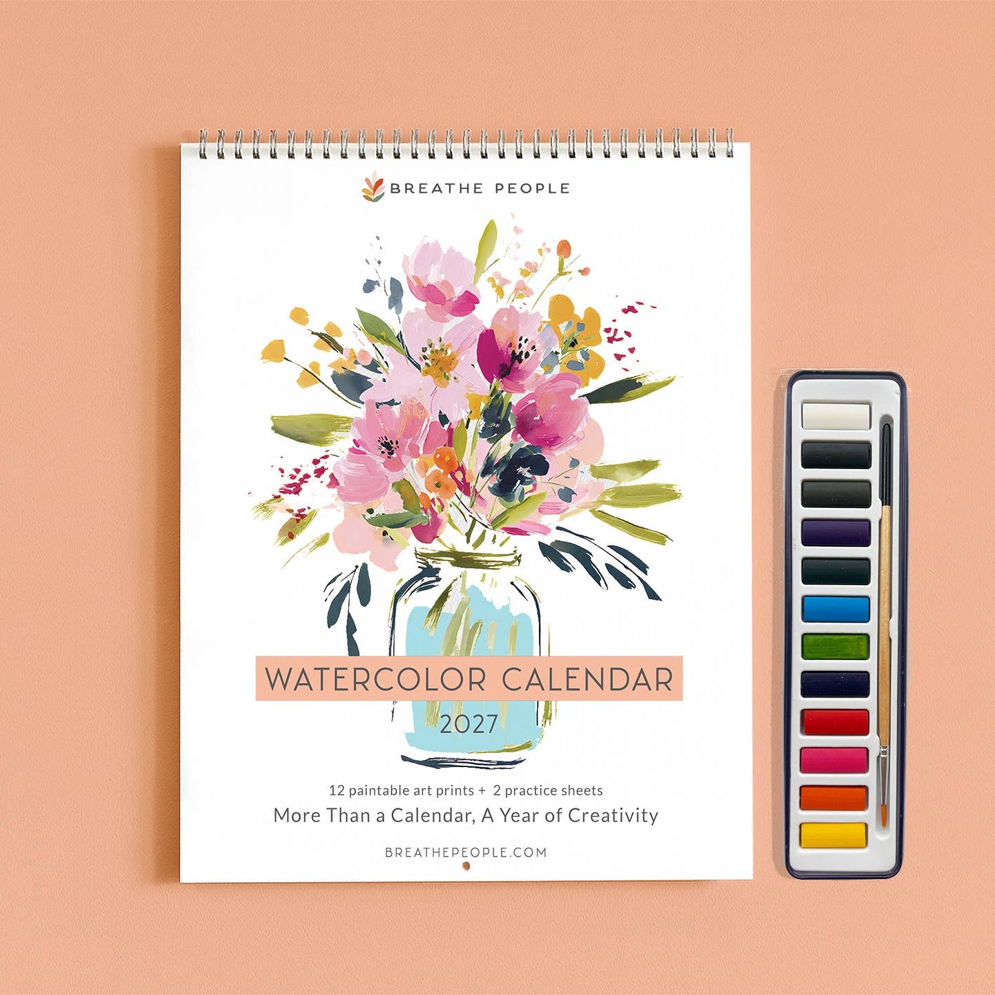A spiral-bound 2027 watercolor art print calendar by Breathe People, featuring a colorful watercolor floral bouquet in a glass jar on the cover, displayed next to a watercolor paint palette on a peach background.